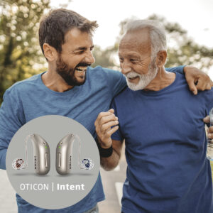 A middle aged man with a beard with his arm around his father, who is wearing oticon hearing aids.