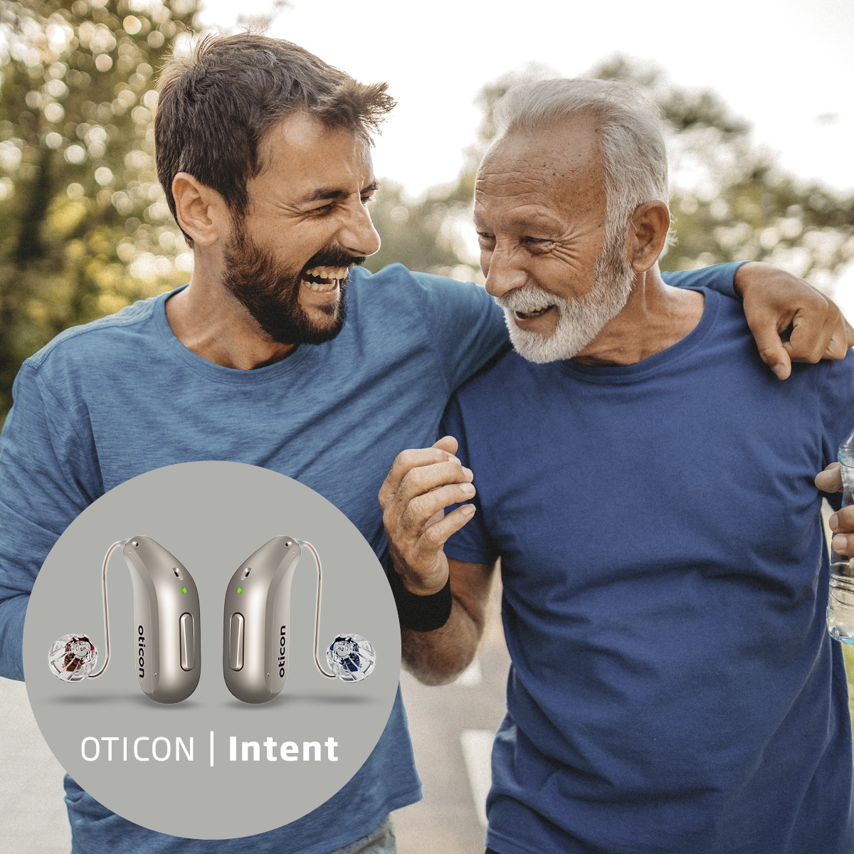 A middle aged man with a beard with his arm around his father, who is wearing oticon hearing aids.