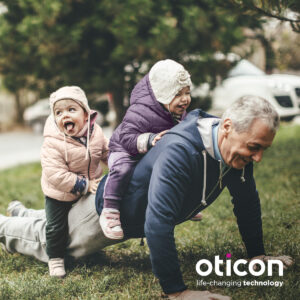 Grandpa doing pushups with toddlers on his back. He's wearing an oticon hearing aid.